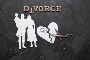 Paper heart cut next to scissors and two rings. Paper couple with a child are next to it. Divorce is written at the top with wooden letters.